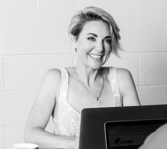 Black and white portrait of a smiling woman at a computer desk wearing a white tank top and pendant necklace