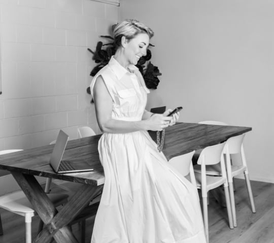 Black and white photo of a woman in a white dress holding a clipboard at a desk with a laptop